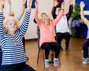 A group of seniors doing a seated exercise class with arms raised up and feet flat on the ground. Seated exercise for people with limited mobility. Propel Physiotherapy.