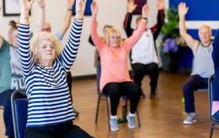 A group of seniors doing a seated exercise class with arms raised up and feet flat on the ground. Seated exercise for people with limited mobility. Propel Physiotherapy.
