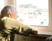 Elderly man stands in the window of his home looking outside longingly. OT psychotherapy Propel Physiotherapy.