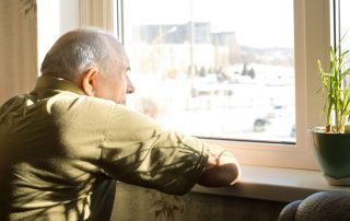 Elderly man stands in the window of his home looking outside longingly. OT psychotherapy Propel Physiotherapy.