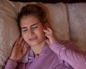 Woman lies in bed holding her jaw and wincing in pain. TMJ disorders management at Propel Physiotherapy.