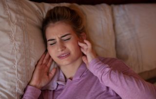 Woman lies in bed holding her jaw and wincing in pain. TMJ disorders management at Propel Physiotherapy.