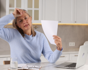 Mature woman fanning herself with piece of paper as she experiences a hot flash while sitting at the kitchen counter working on her laptop. Most common menopause symptoms and pelvic floor physiotherapy at Propel Physiotherapy.