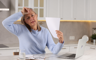 Mature woman fanning herself with piece of paper as she experiences a hot flash while sitting at the kitchen counter working on her laptop. Most common menopause symptoms and pelvic floor physiotherapy at Propel Physiotherapy.
