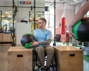 Man in wheelchair in a gym working with a big medicine ball between two large wooden blocks with his physiotherapist watching him. COM-B framework behaviour change science tool for physiotherapy. Propel Physiotherapy
