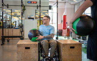 Man in wheelchair in a gym working with a big medicine ball between two large wooden blocks with his physiotherapist watching him. COM-B framework behaviour change science tool for physiotherapy. Propel Physiotherapy