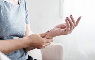 Middle-age female sitting on the couch at home holding her left forearm with her right hand with left hand palm facing up. She is experiencing menopause joint and muscle pain, carpal tunnel. Propel Physiotherapy can effectively help manage menopause joint and muscle pain and other MSK symptoms by focusing on exercises that strengthen muscles, improve flexibility, and enhance joint mobility, as well as providing education, manual therapy and other modalities that aid in pain management.