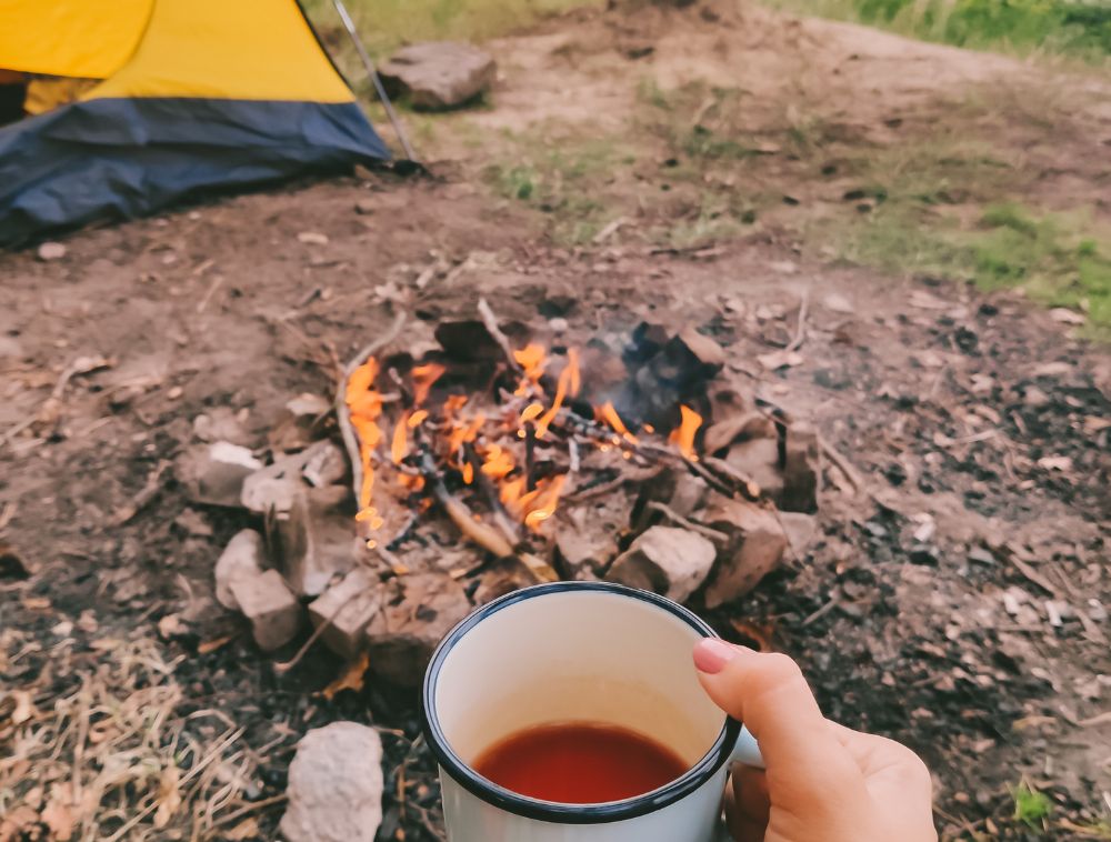Woman's hand holding a cup of coffee in front of a campfire and a tent. Brianna James Physiotherapy Assistant Occupational Therapy Propel Physiotherapy Peterborough enjoys camping.