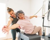 Mature woman holds onto bar onto trapeze bar with left hand while seated and reaching back with other hand under the supervision of her physical therapist. Dual task training - a cognitive-motor approach to fall prevention and balance. Propel Physiotherapy.