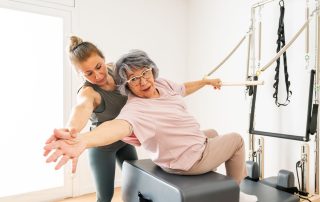 Mature woman holds onto bar onto trapeze bar with left hand while seated and reaching back with other hand under the supervision of her physical therapist. Dual task training - a cognitive-motor approach to fall prevention and balance. Propel Physiotherapy.