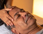 Older man lying on massage table with hands of massage therapists on temples. The benefits of massage therapy for headaches at Propel Physiotherapy.