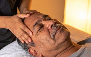 Older man lying on massage table with hands of massage therapists on temples. The benefits of massage therapy for headaches at Propel Physiotherapy.