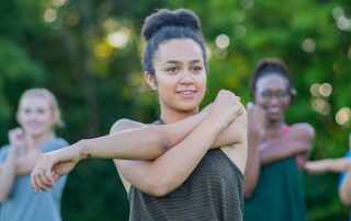 Group of people in a park holding their left arm across the front of the chest with the right arm to stretch their left shoulder. Types of Stretching: Static, Dynamic, Ballistic and PNF Stretching Explained by Kathy Mileski at Propel Physiotherapy.